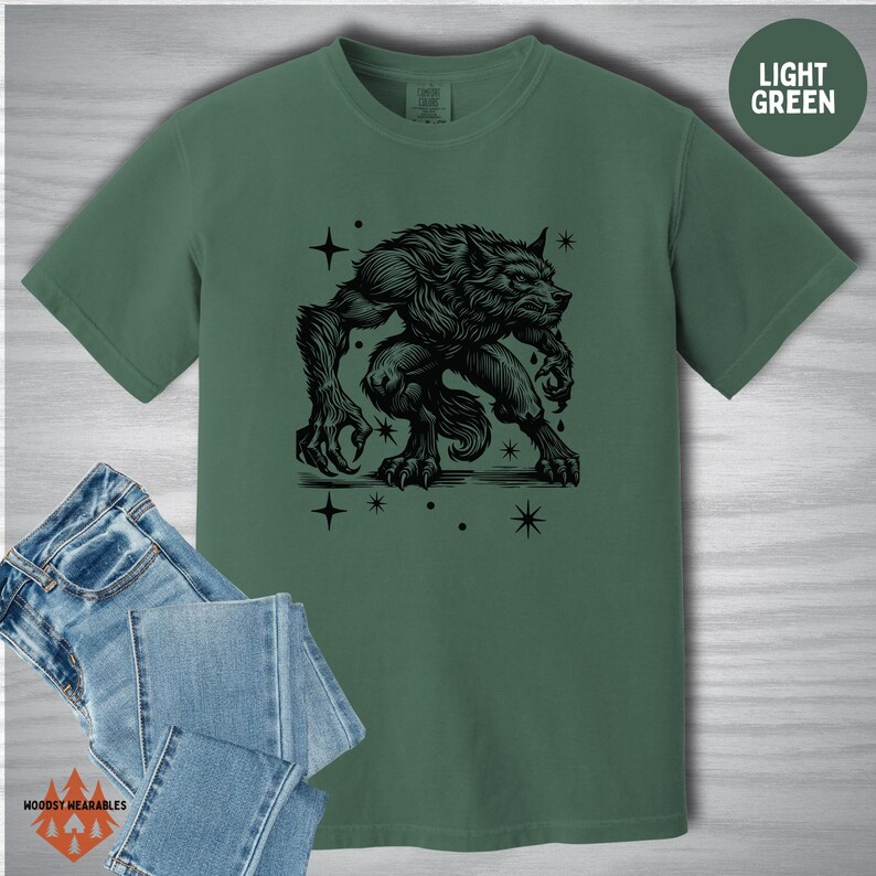 Flat lay of a Comfort Colors shirt in light green featuring a black werewolf design in linocut style. The werewolf crouches surrounded by stars, printed alongside a folded pair of jeans. “LIGHT GREEN” label in the upper right.