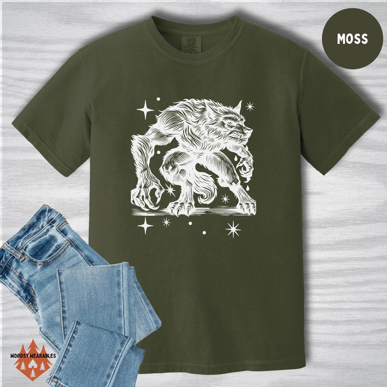 Flat lay of a Comfort Colors t-shirt in moss green with folded jeans. The shirt features a white linocut-style illustration of a crouching werewolf surrounded by stars. The werewolf has exaggerated claws, fur texture, and a crouched stance. “MOSS” label in top right corner.