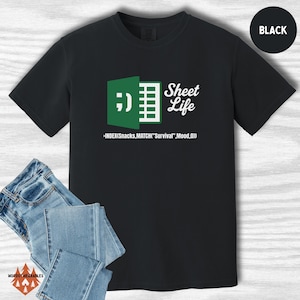 May include: Black t-shirt featuring a green and white spreadsheet graphic with the words "Sheet Life." The design includes a smiley face and a formula. Blue jeans are visible in the foreground.