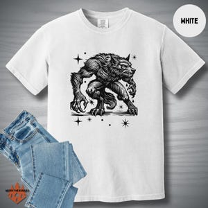Flat lay of a white Comfort Colors tee featuring a black linocut-style werewolf design with a vintage horror aesthetic. Styled with light denim jeans. "WHITE" label in the top right corner.