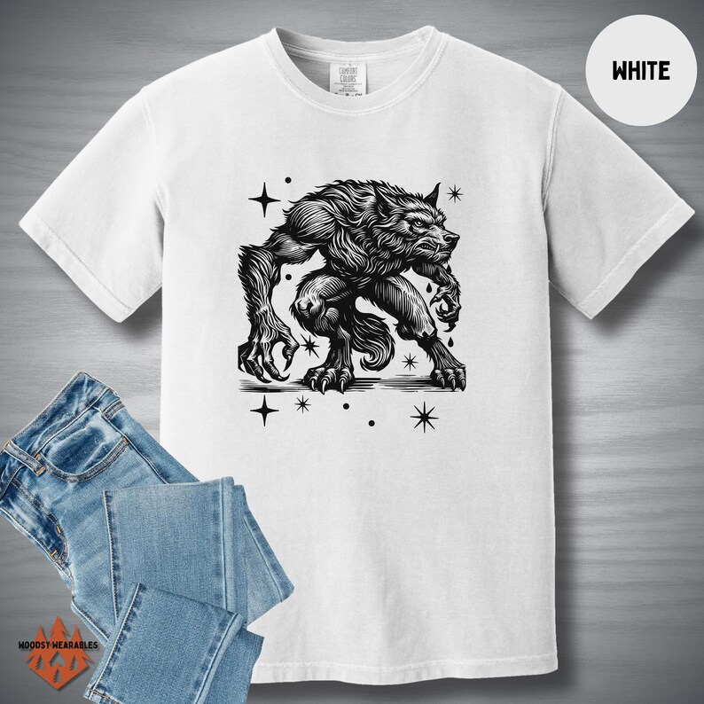 Flat lay of a white Comfort Colors tee featuring a black linocut-style werewolf design with a vintage horror aesthetic. Styled with light denim jeans. "WHITE" label in the top right corner.