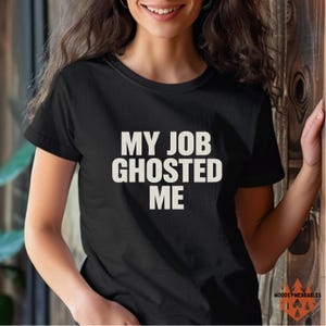 May include: Black crew neck t-shirt with the text "MY JOB GHOSTED ME" in white block letters. The shirt is a short-sleeved tee. The bottom right corner of the shirt has a logo that says "WOODSY-WEARABLES".