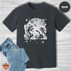 Flat lay of a Comfort Colors t-shirt in denim blue with a white werewolf design in a vintage, carved linocut style. The creature is mid-snarl, surrounded by stars. Includes folded jeans and a “DENIM” label in the upper right.