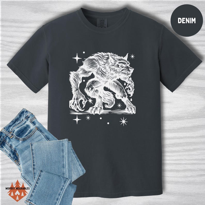 Flat lay of a Comfort Colors t-shirt in denim blue with a white werewolf design in a vintage, carved linocut style. The creature is mid-snarl, surrounded by stars. Includes folded jeans and a “DENIM” label in the upper right.