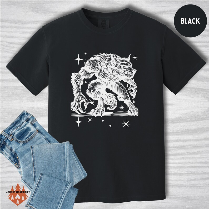 Black Comfort Colors shirt featuring a white vintage-style werewolf illustration. The werewolf appears fierce and beastly, surrounded by scattered stars. Flat lay with denim jeans. “BLACK” label in the top right corner.