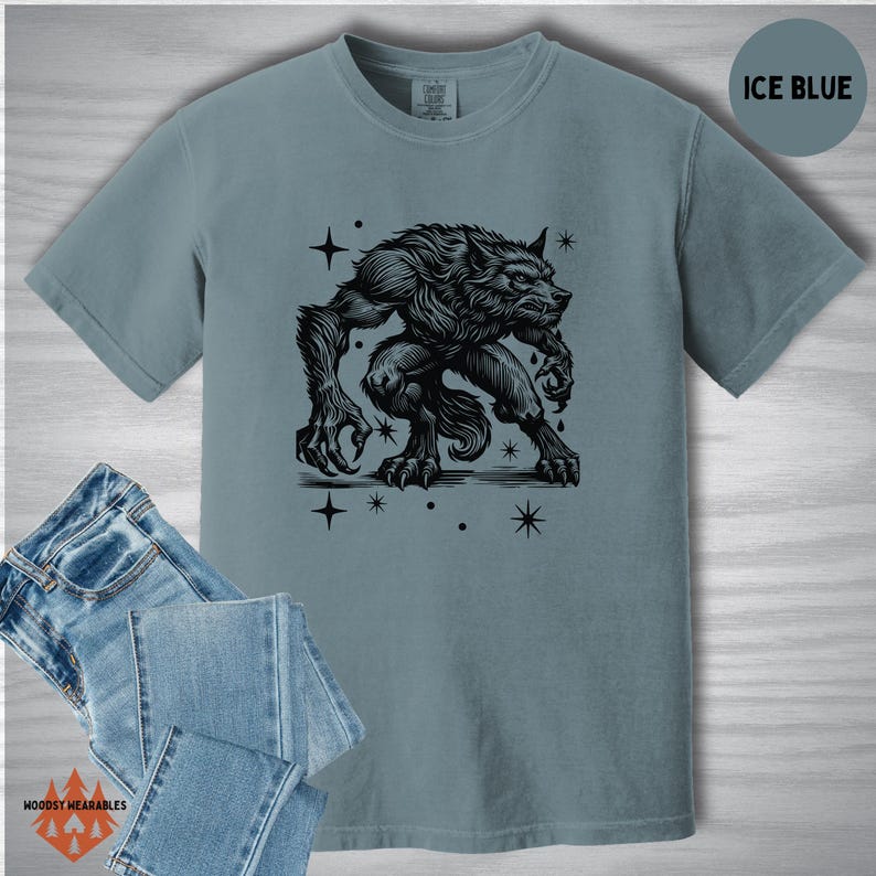 Comfort Colors t-shirt in ice blue with a black linocut-style werewolf graphic and cosmic star elements. Flat lay styling includes denim jeans. “ICE BLUE” label in the upper right.