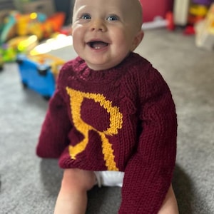 May include: A baby wearing a maroon knitted sweater with a yellow letter "R" on the front. The baby is smiling and looking to the side.