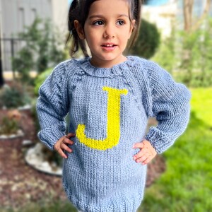 May include: A light blue knitted sweater with a yellow letter J on the front. The sweater is worn by a young child.