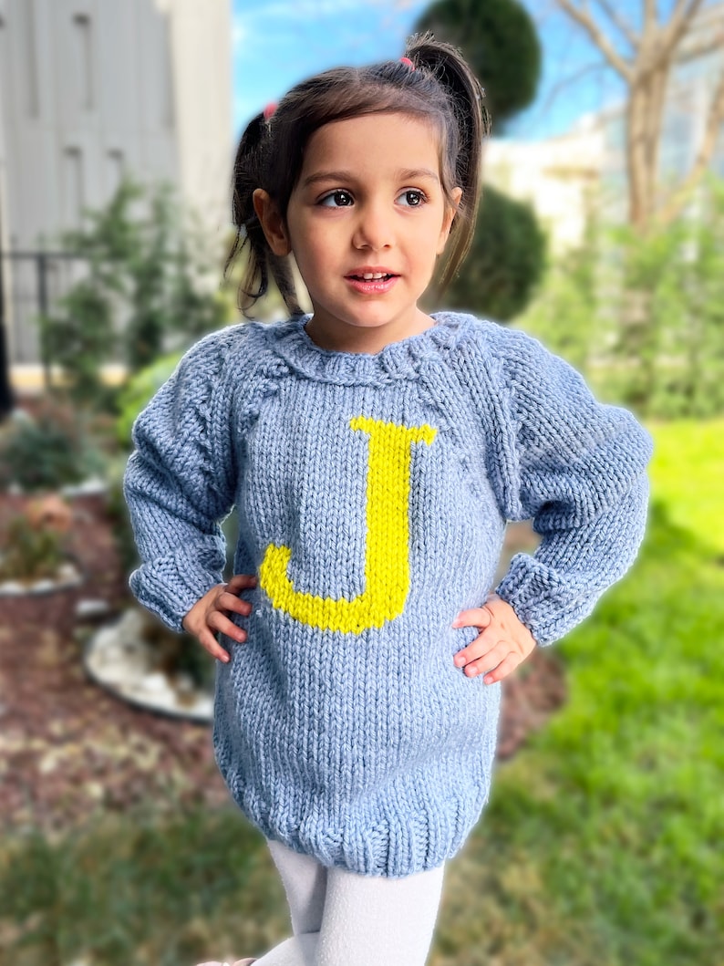 May include: A light blue knitted sweater with a yellow letter J on the front. The sweater is worn by a young child.