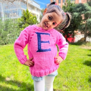 May include: A young girl wearing a pink knitted sweater with a blue letter "E" on the front. She is standing in a grassy area with a building in the background.