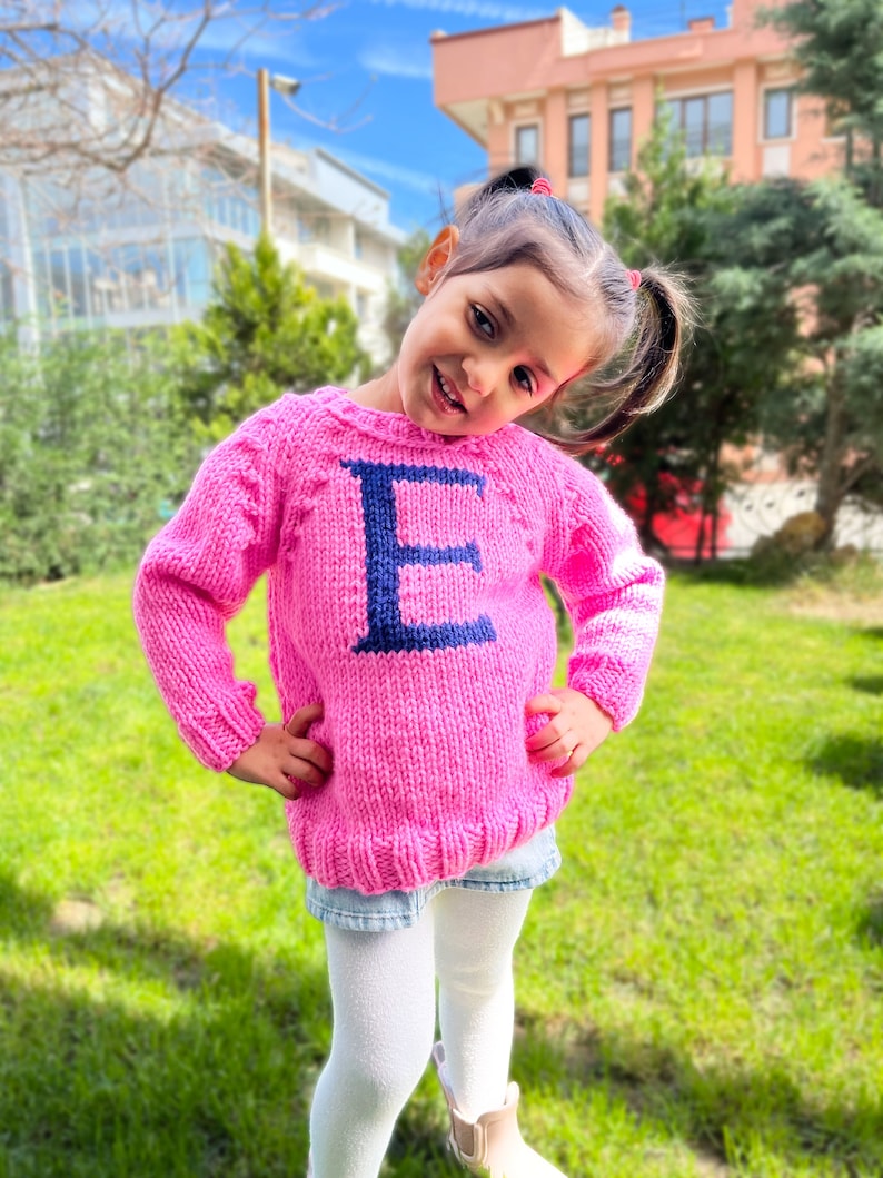 May include: A young girl wearing a pink knitted sweater with a blue letter "E" on the front. She is standing in a grassy area with a building in the background.