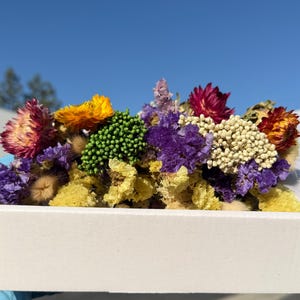 May include: A white rectangular cardboard box filled with a variety of dried flowers including purple statice, yellow yarrow, bright orange strawflowers, deep red strawflowers, green flower buds, and small white clusters of achillea millefolium.