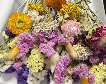 May include: A vibrant bouquet of dried flowers in shades of yellow, orange, purple, pink, and white. The arrangement includes various textures and shapes, with a focus on the colorful blooms and delicate stems. The flowers are arranged in a white container.