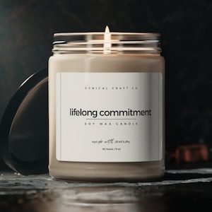 May include: A soy wax candle in a clear glass jar with a white label that reads "CYNICAL CRAFT CO lifelong commitment SOY WAX CANDLE made with sarcasm 50 hours/9 oz".
