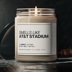 May include: A brown glass candle jar with a white label that reads "Smells Like AT&T Stadium A Cowboys Victory Never Smelt Better Hand Poured in California 9 oz 60 Hours | Soy Wax 1 AT&T Way, Arlington, TX 76011".