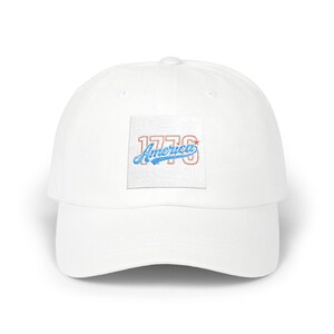 Puede incluir: Gorra de béisbol blanca con visera curva. La gorra presenta un parche cuadrado con el texto "1776 America" bordado en rojo y azul. El sombrero está hecho de un material suave y tiene un estilo clásico e informal.