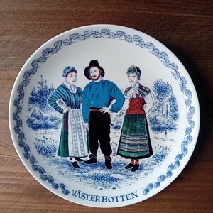 May include: White ceramic plate with a blue and white border featuring a decorative leaf pattern. The plate depicts three figures in traditional clothing, with the text "VÄSTERBOTTEN" at the bottom. The figures are set against a blue and white landscape.