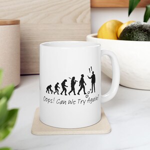 May include: White ceramic mug featuring a black evolution of man graphic. The design includes figures progressing from ape-like to a modern human, with the text "Oops! Can We Try Again?" printed below the figures.