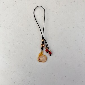K&ouml;nnte beinhalten: Ein goldfarbener Hasenanh&auml;nger mit einem rosafarbenen, wei&szlig;en K&ouml;rper und einem roten Punktauge h&auml;ngt an einer schwarzen Kordel. Der Anh&auml;nger ist mit goldenen und roten Perlen verziert. Der Anh&auml;nger ist wahrscheinlich ein Anh&auml;nger oder Schl&uuml;sselanh&auml;nger.