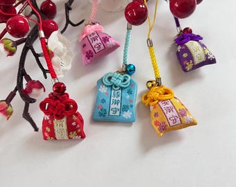 May include: A collection of colorful Japanese omamori charms. The charms are in various colors, including red, pink, blue, yellow, and purple. Each charm has a different design and is decorated with floral patterns and Japanese characters. They are hanging from a branch with red berries.