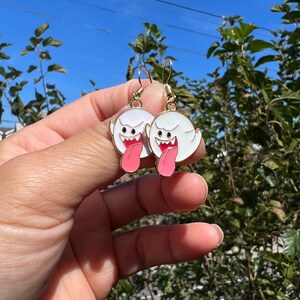 May include: A pair of gold-colored hook earrings featuring white ghost-shaped charms with black eyes, red mouths, and pink tongues. The earrings are held up against a backdrop of green foliage and a blue sky.