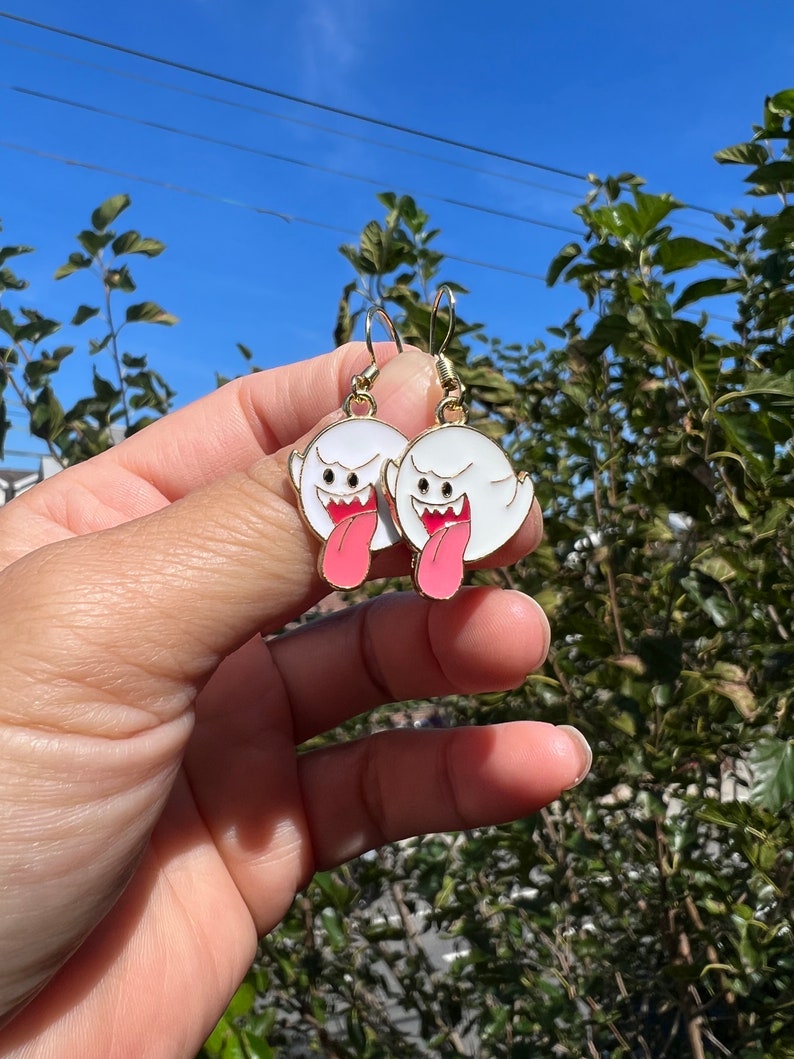 May include: A pair of gold-colored hook earrings featuring white ghost-shaped charms with black eyes, red mouths, and pink tongues. The earrings are held up against a backdrop of green foliage and a blue sky.