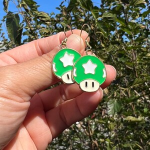 May include: A pair of green and white mushroom earrings with gold-colored hooks. Each earring features a green mushroom cap with a white star and a white base. The earrings are held up against a background of green foliage.