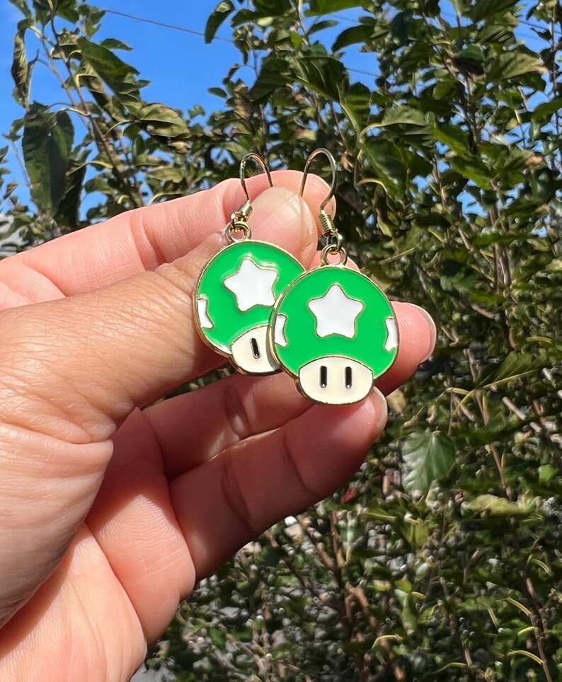 May include: A pair of green and white mushroom earrings with gold-colored hooks. Each earring features a green mushroom cap with a white star and a white base. The earrings are held up against a background of green foliage.