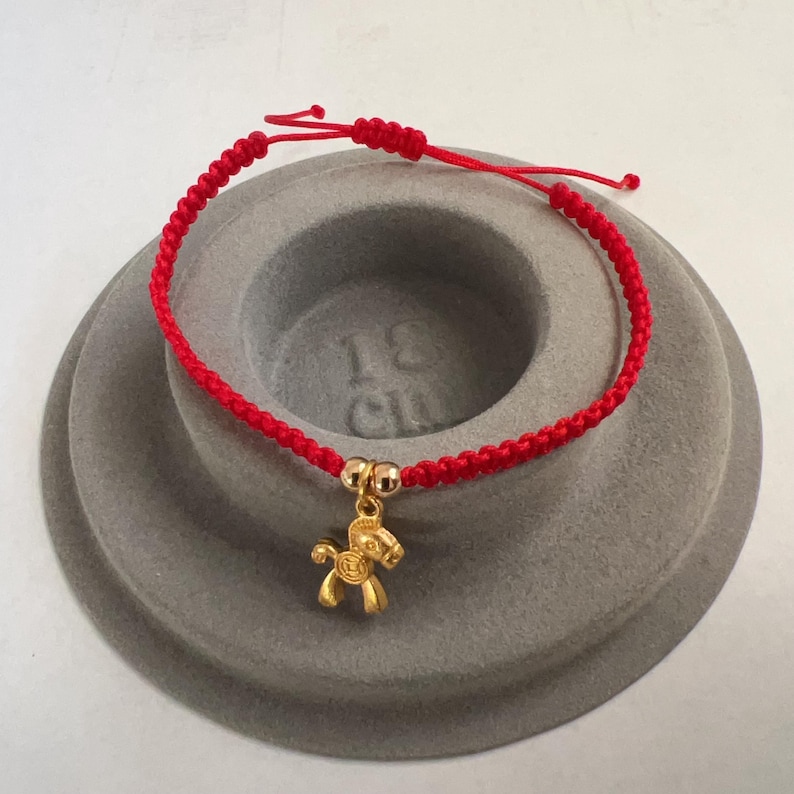 May include: Red braided bracelet with a gold-colored horse charm. The bracelet is adjustable with sliding knots. The horse charm has detailed features. The bracelet is displayed on a gray, tiered surface.