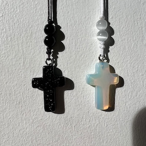 May include: Two necklaces with cross pendants. One necklace has a black cross pendant with black beads and a black cord. The other necklace has a white cross pendant with white beads and a white cord.