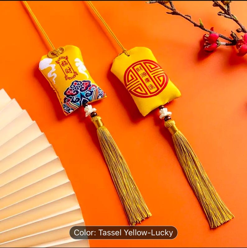 May include: Two yellow lucky charm pendants with gold tassels. Each pendant has a different design, one with Chinese characters and the other with a circular emblem. The pendants are suspended against an orange backdrop.