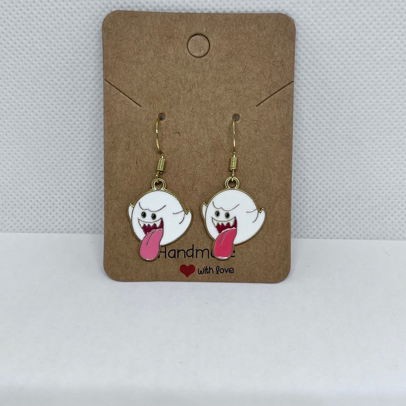 May include: A pair of gold-colored hook earrings featuring a white ghost design with a pink tongue sticking out. The earrings are attached to a brown card with the words "Handmade with love".