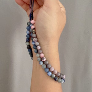 May include: Two beaded bracelets, one with blue and gray stones and the other with pink and gray stones, are tied together with a dark blue cord.