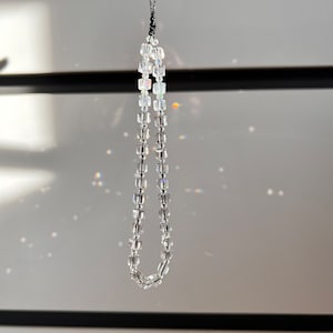 May include: A clear crystal beaded necklace with a black cord. The necklace is hanging from a white surface.