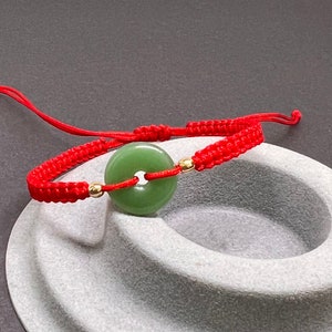 May include: A red string bracelet with a green jade circle charm. The charm has a gold ring in the center.