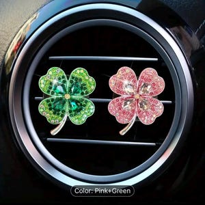 May include: Two car air vent clips shaped like four-leaf clovers. One clover is green with clear rhinestones and the other is pink with clear rhinestones.  The text "Color: Pink+Green" is visible below the clovers.