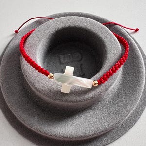 May include: A red braided cord bracelet featuring a white cross pendant with iridescent accents. The cross is flanked by small gold-colored beads. The bracelet is displayed on a gray, circular surface.