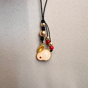 K&ouml;nnte beinhalten: Eine goldfarbene Anh&auml;ngerkette mit einem wei&szlig;en Hasenanh&auml;nger und einem roten Akzent. Die Kette hat goldene und rote Perlen, die auf einer schwarzen Kordel aufgef&auml;delt sind. Der Hasenanh&auml;nger ist mit goldenen Akzenten und einem kleinen roten Punkt versehen.