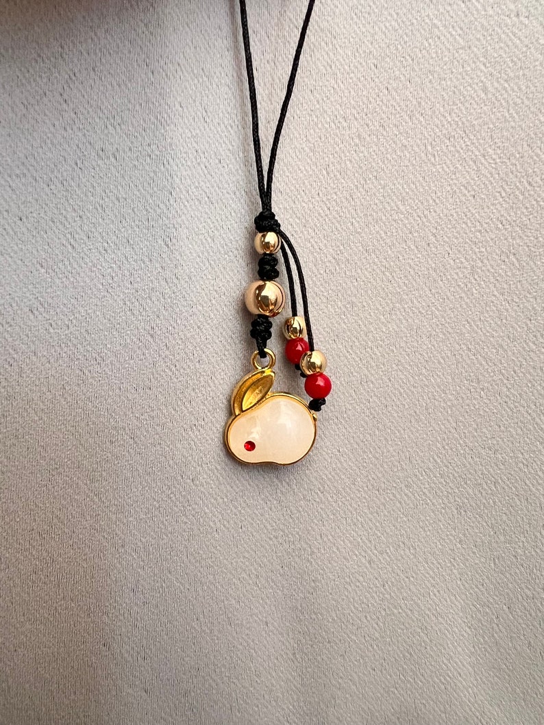 K&ouml;nnte beinhalten: Eine goldfarbene Anh&auml;ngerkette mit einem wei&szlig;en Hasenanh&auml;nger und einem roten Akzent. Die Kette hat goldene und rote Perlen, die auf einer schwarzen Kordel aufgef&auml;delt sind. Der Hasenanh&auml;nger ist mit goldenen Akzenten und einem kleinen roten Punkt versehen.