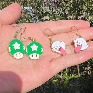 May include: A pair of gold-toned earrings featuring green mushroom charms with white star details and a pair of white ghost charms with pink tongues. The earrings are designed with a gold hook.