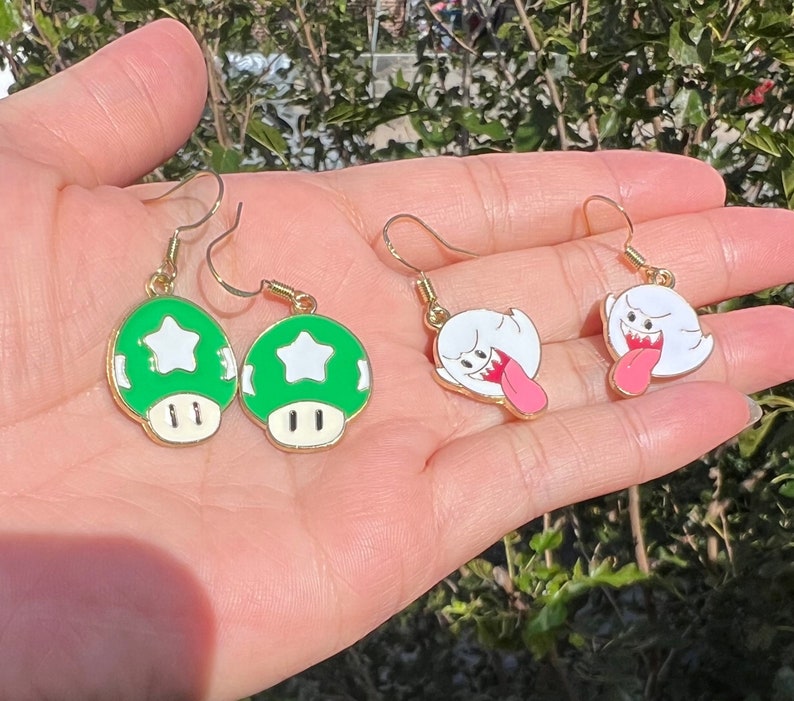 May include: A pair of gold-toned earrings featuring green mushroom charms with white star details and a pair of white ghost charms with pink tongues. The earrings are designed with a gold hook.