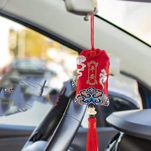 May include: Red car charm with Chinese characters and a decorative tassel. The charm is rectangular, with ornate patterns in gold, blue, and white. It hangs from a red string, suspended in front of a car's interior.