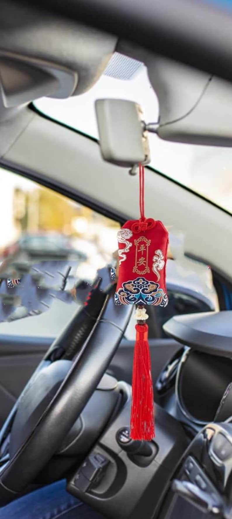 May include: Red car charm with Chinese characters and a decorative tassel. The charm is rectangular, with ornate patterns in gold, blue, and white. It hangs from a red string, suspended in front of a car's interior.