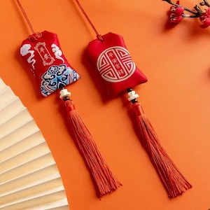 May include: Two red fabric pouches with tassels hang against an orange background. One pouch has embroidered Chinese characters and a cloud design. The other has a circular design with Chinese characters. Both have long orange tassels.