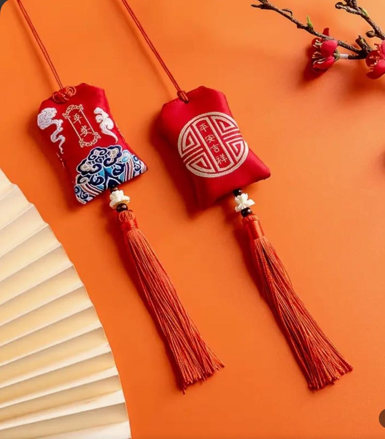May include: Two red fabric pouches with tassels hang against an orange background. One pouch has embroidered Chinese characters and a cloud design. The other has a circular design with Chinese characters. Both have long orange tassels.