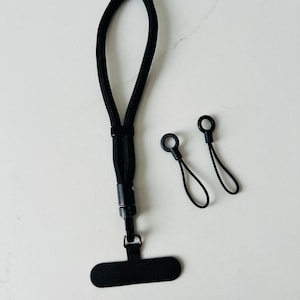 May include: A black wrist lanyard featuring a woven cord, a plastic connector, and a flat T-shaped attachment, accompanied by two small black loop tethers with circular plastic ends, designed for securing small electronic devices or accessories.