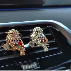 May include: Two gold-tone bird car air freshener ornaments with colorful rhinestones. The birds are perched on a branch and have a clip on the back to attach to a car vent.