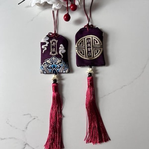 May include: Two purple good luck charms with embroidered designs and long red tassels. One charm features a cloud and wave pattern, the other a geometric design. The charms hang from red cords.