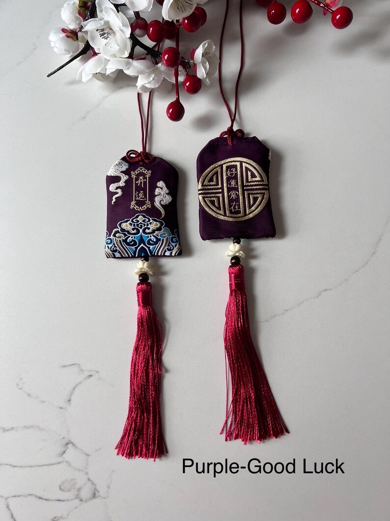 May include: Two purple good luck charms with embroidered designs and long red tassels. One charm features a cloud and wave pattern, the other a geometric design. The charms hang from red cords.