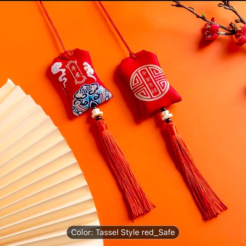 May include: Two red fabric ornaments with tassels. One features embroidered blue and white designs and Chinese characters. The other has a circular design with Chinese characters. Both have red cords for hanging.
